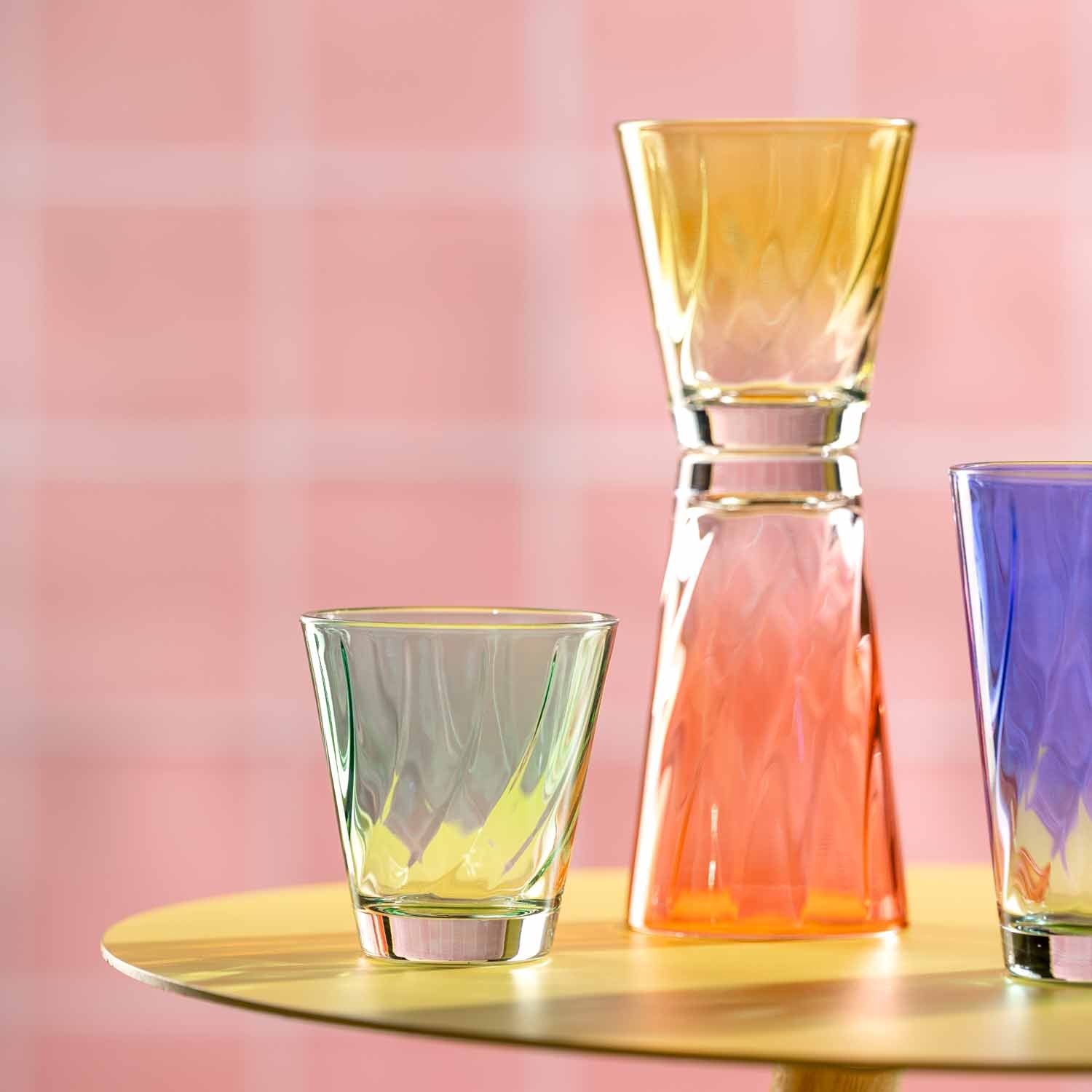Trinkgläser TWIST mit farbigen Flüssigkeiten auf goldenem Tablett, rosa Hintergrund