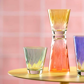 Trinkgläser TWIST mit farbigen Flüssigkeiten auf goldenem Tablett, rosa Hintergrund