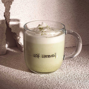 Tasse EMOZIONE mit Matcha-Getränk, Gravur „…ich liebs!", Sonnenlicht, heller Untergrund