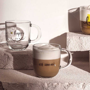 Tasse EMOZIONE mit Schriftzug, gefüllt mit Kaffee, neben leerer Tasse auf Textilien