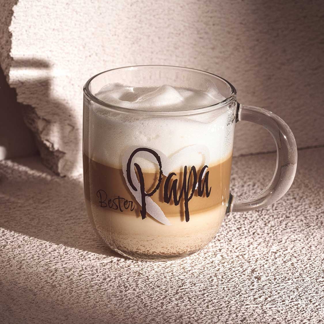 Tasse EMOZIONE mit "Bester Papa" Gravur, gefüllt mit Kaffee und Milch, Sonnenlicht auf Teppich