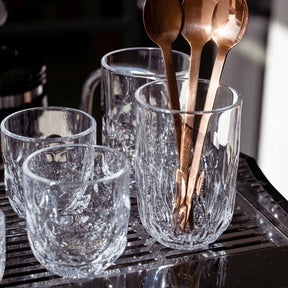 Latte Macchiato Glas GOCCE mit Wasser und Kupferlöffeln auf schwarzem Tablett