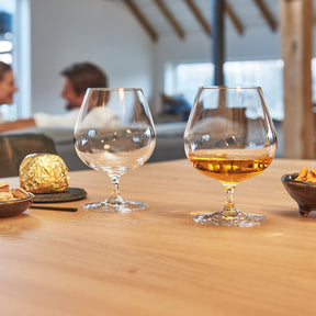 Cognacschwenker CIAO+ gefüllt und leer auf Holztisch mit Snacks und Personen im Hintergrund