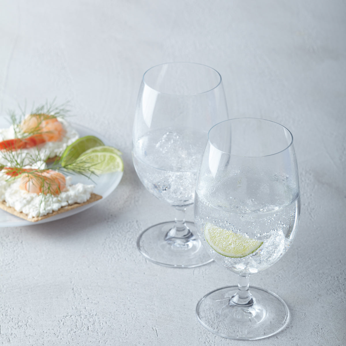 Zwei Wassergläser DAILY mit Eis und Limette, daneben Appetithäppchen auf Teller