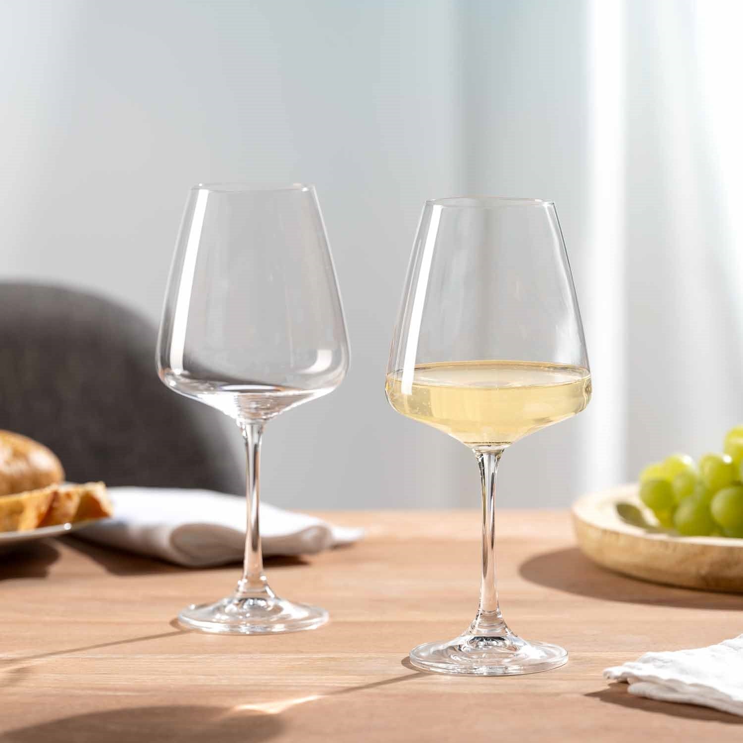 Zwei Weißweingläser PALADINO auf Holztisch, eines leer, eines mit Weißwein gefüllt, neben Obst und Servietten