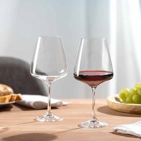 Zwei Rotweingläser PALADINO auf Holztisch, eines leer, eines mit Rotwein gefüllt, daneben Obst und Gebäck