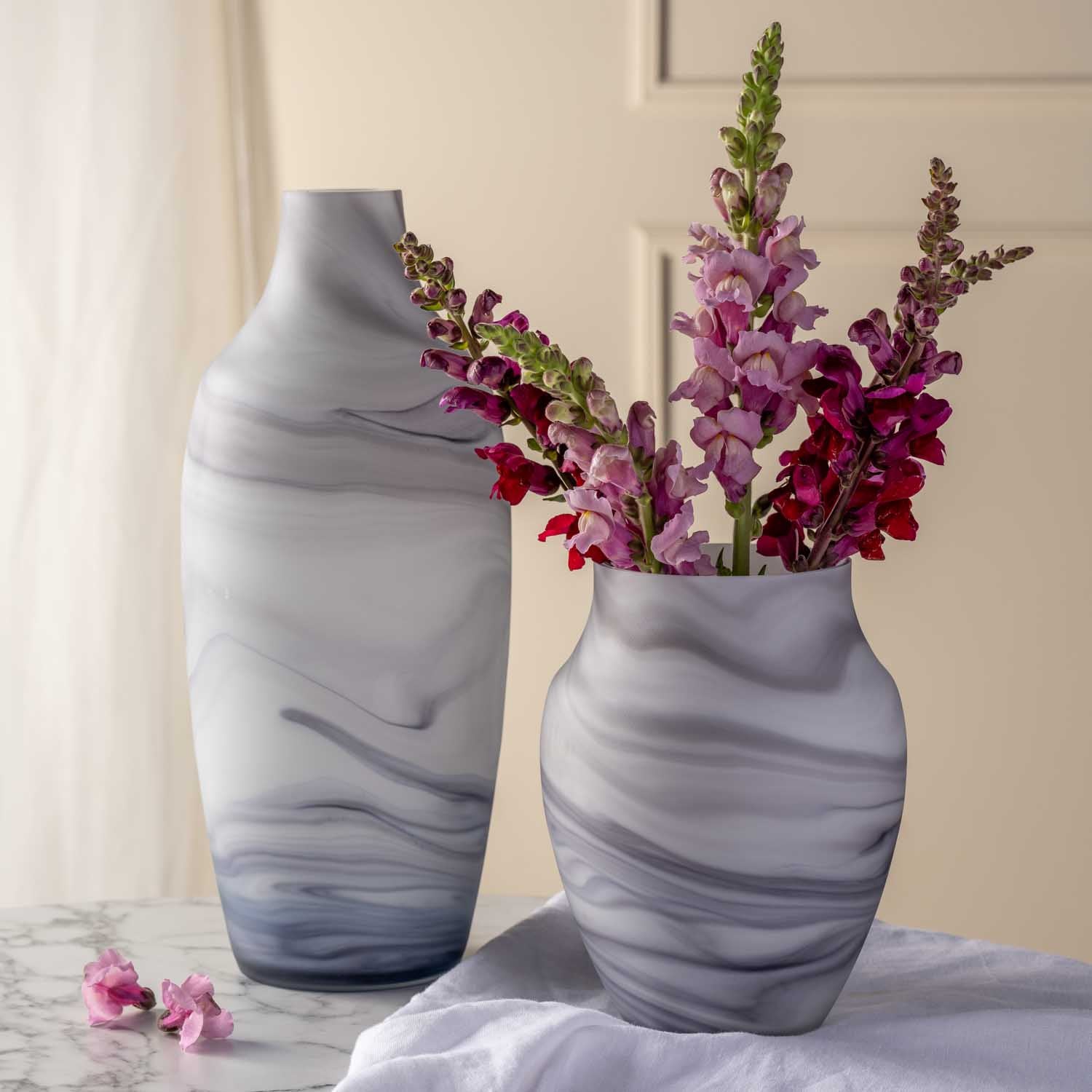 Vase POESIA mit Marmoroptik, gefüllt mit violetten und roten Blüten, neben einer zweiten Vase auf Marmortisch