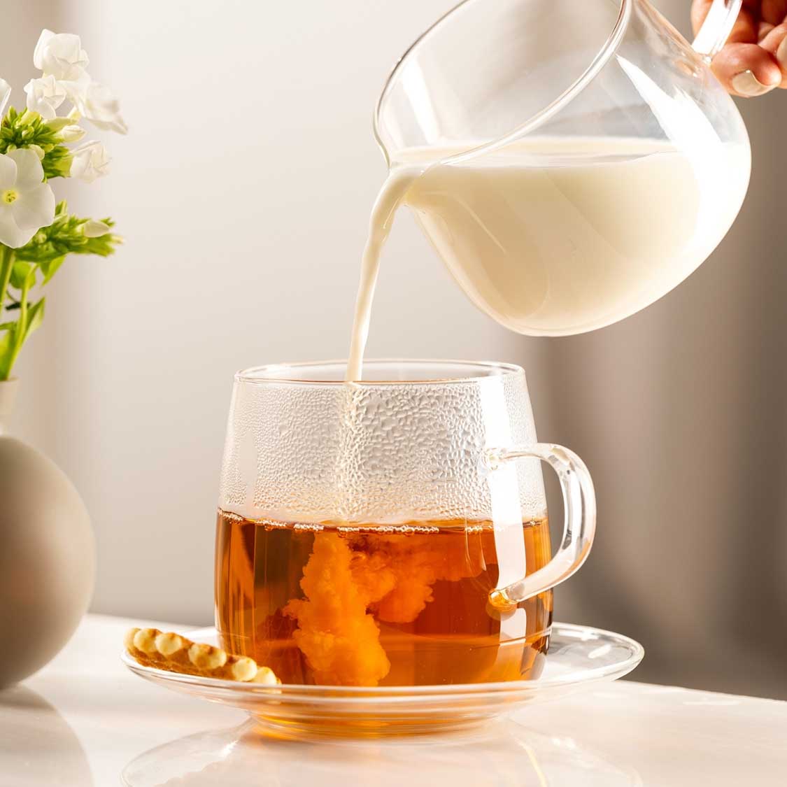 Tee wird in Glastasse mit Untertasse eingegossen, Blumen daneben