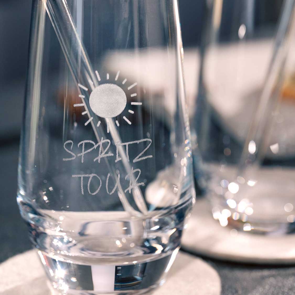 Trinkglas mit Sonne und "SPRITZ TOUR" Gravur auf gedecktem Tisch