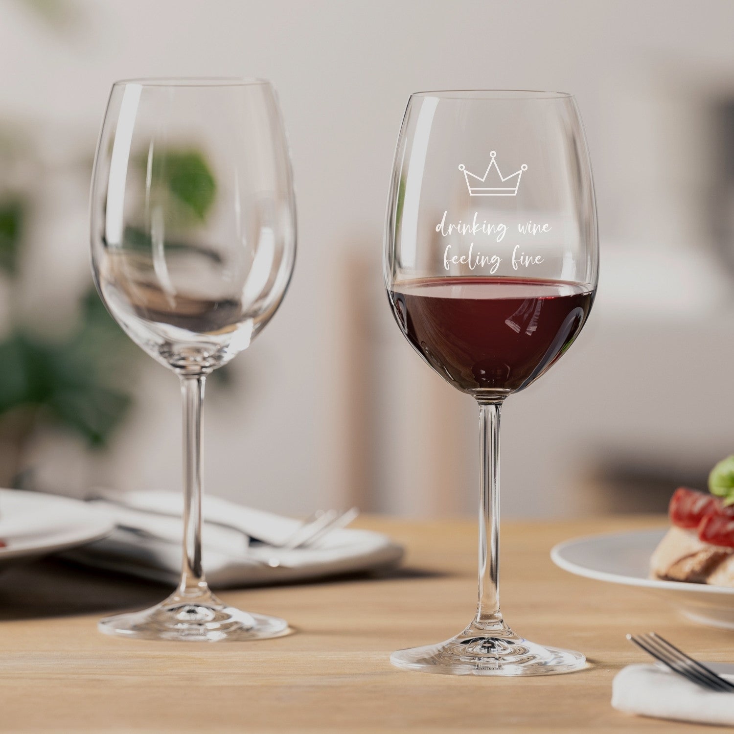 Zwei Rotweingläser auf gedecktem Tisch, eines mit Gravur "drinking wine feeling fine