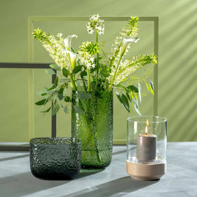 Windlicht LEGNO mit Holzsockel neben Glasvasen mit grünen Blüten auf Tisch