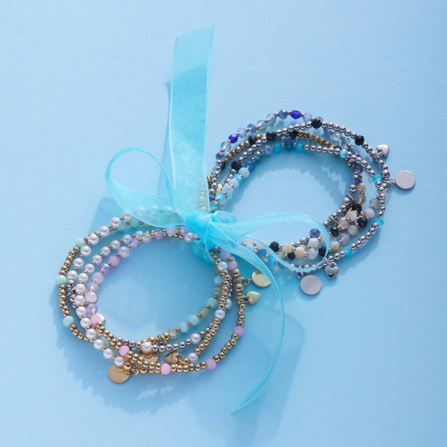 Zwei mehrfarbige Armbänder UNITA You & Me CIAO mit Perlen und Charms auf hellblauem Hintergrund