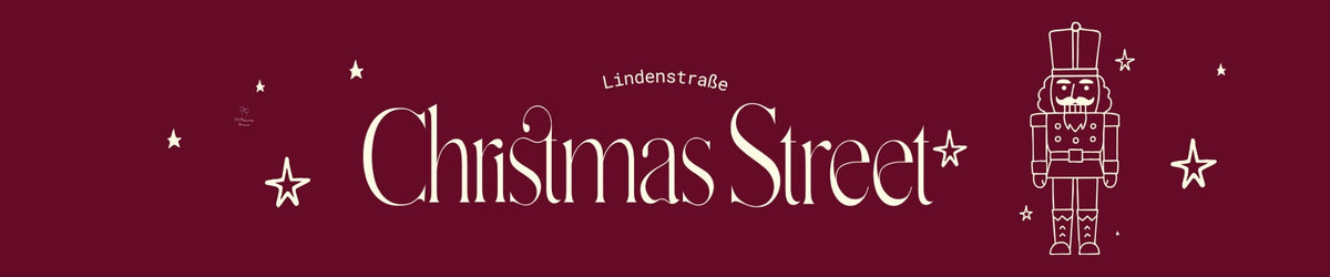 Breites Banner mit burgunderrotem Hintergrund. Zentral steht der Schriftzug ‚Christmas Street‘ in dekorativer, cremefarbener Schrift, darüber der kleine Text ‚Lindenstraße‘. Links und rechts sind mehrere gezeichnete Sterne verteilt. Rechts neben dem Schriftzug ist eine stilisierte cremefarbene Nussknacker-Figur dargestellt.