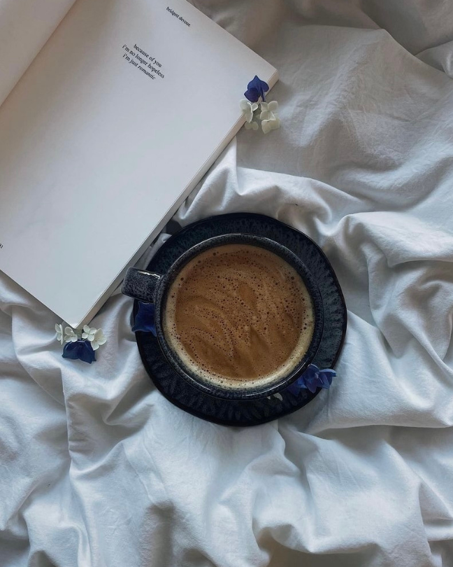 Eine dunkelblaue Keramiktasse mit Unterteller steht inmitten eines Bettes auf weißer Bettwäsche neben einem aufgeschlagenen Buch und dekorativen Blüten.