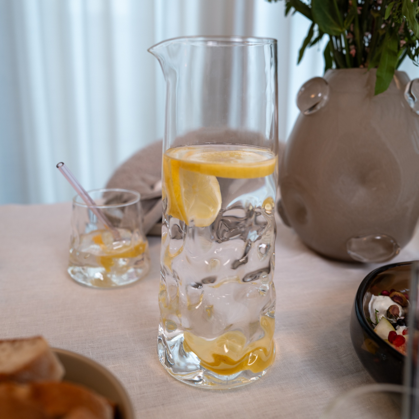Transparente Glaskaraffe mit Zitronenscheiben und Wasser auf einem gedeckten Tisch, im Hintergrund eine Vase und ein Trinkglas.