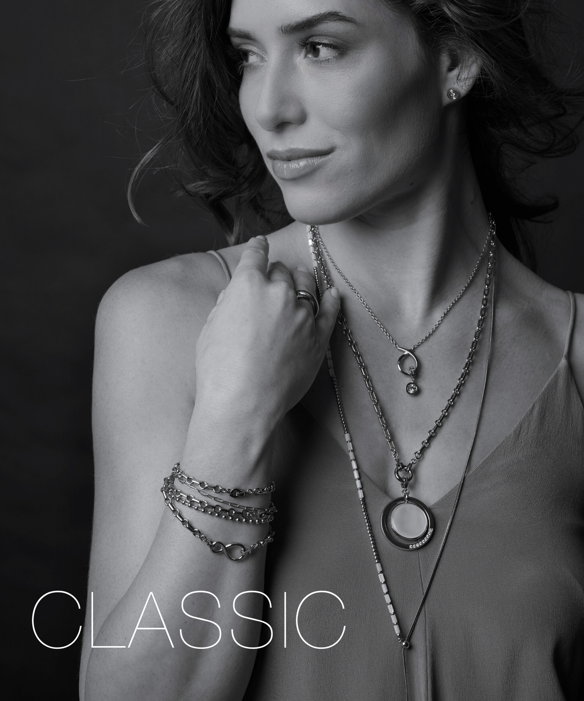 Eine Frau mit dunklem Haar blickt nachdenklich zur Seite und trägt mehrere feine Ketten und Armbänder. Der zeitlose Schmuck unterstreicht ihren eleganten und ruhigen Ausdruck.