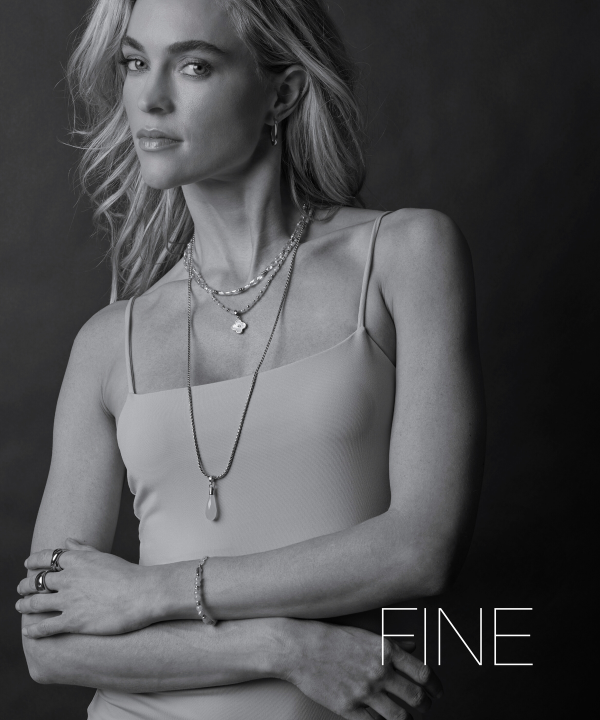 Schwarz-weiß-Foto einer Frau im Trägertop mit feinen Ketten und Armbändern. Sie verschränkt die Arme. Schriftzug ‚Fine‘ im Bild.