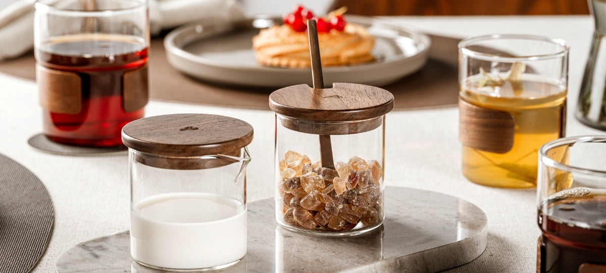 Zwei Glasbehälter mit Deckeln aus dunklem Holz, gefüllt mit Milch und braunem Kandiszucker, stehen auf einer Marmorplatte, umgeben von Teegläsern mit Holzmanschette und einem Dessert im Hintergrund.