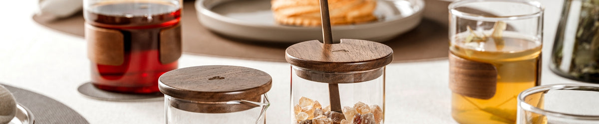 Zwei Glasbehälter mit dunklen Holzdeckeln, einer mit Milch und einer mit braunem Kandiszucker samt Löffel, arrangiert zwischen Teegläsern mit Holzmanschette und einem Dessert im Hintergrund.