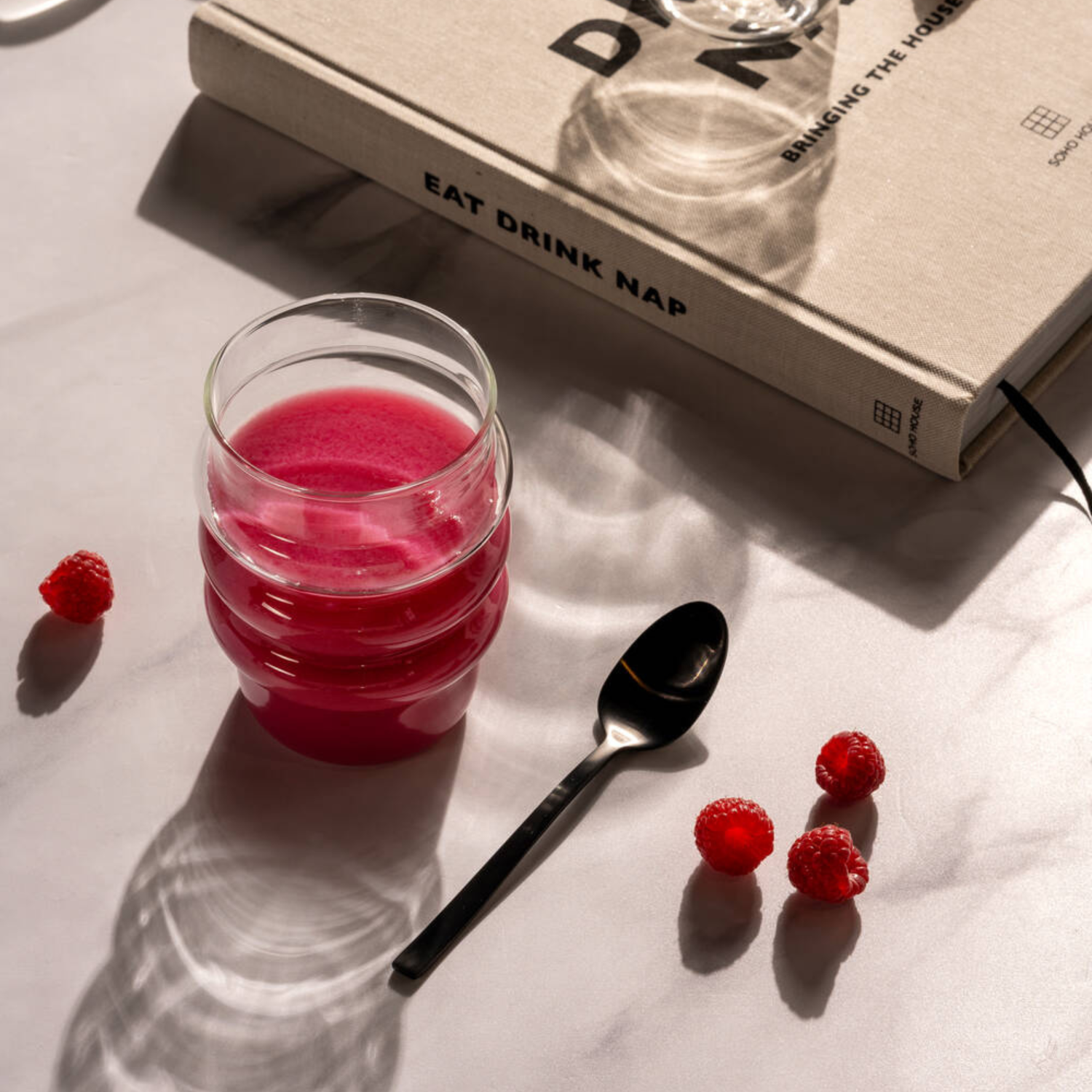 Ein Glas im Bubble Design mit intensiv pinkem Himbeer-Smoothie steht neben einem Löffel und mehreren frischen Himbeeren auf hellem Untergrund mit Lichtreflexen.