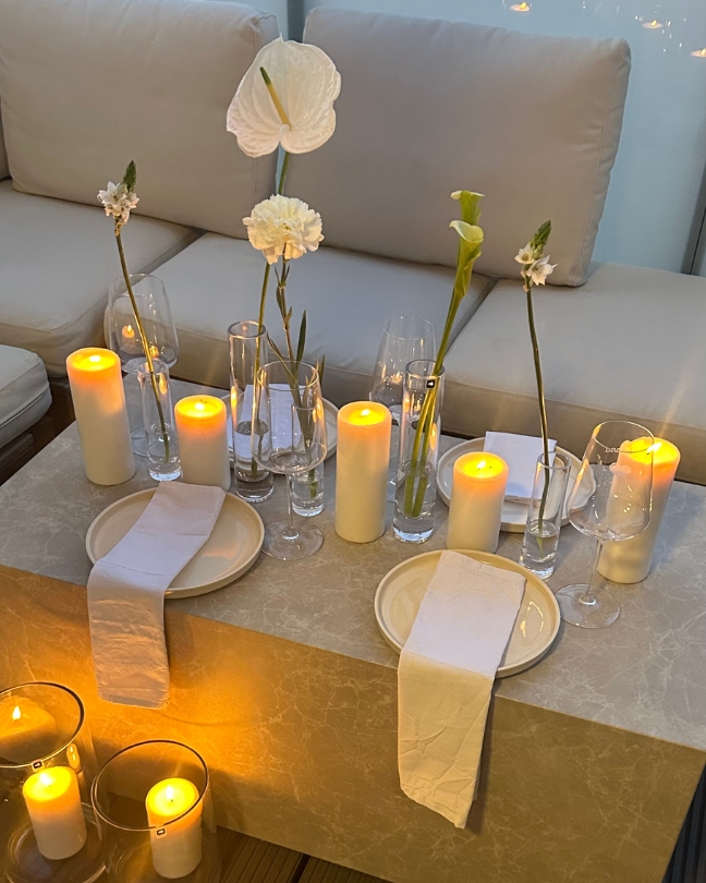 Ein stilvoll gedeckter Tisch mit cremefarbenem Geschirr, weißen Stoffservietten, LED-Stumpenkerzen und schlanken Glasvasen mit einzelnen weißen Blumen schafft eine elegante, stimmungsvolle Atmosphäre auf einer Lounge-Terrasse.