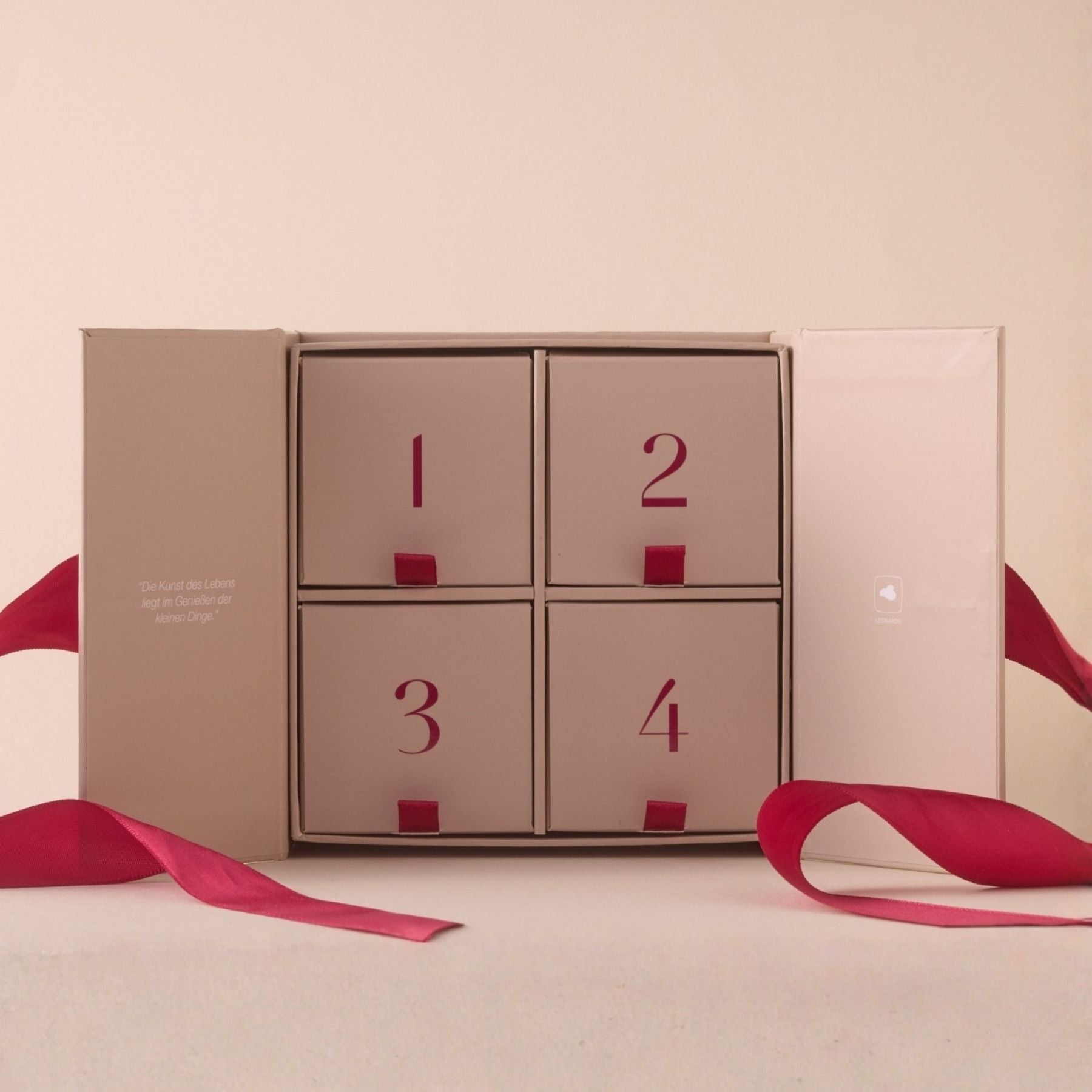 Geöffneter LEONARDO Schmuck Adventskalender mit vier nummerierten Fächern, roten Seidenbändern, beige-rosafarbener Verpackung auf hellem Hintergrund
